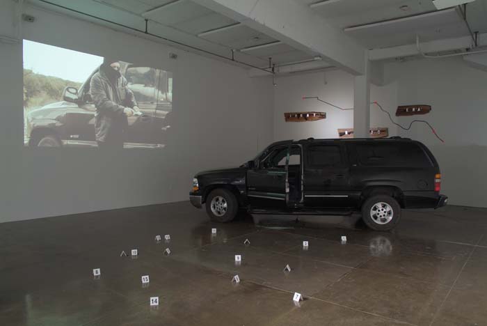 Installation with a black Suburban, crime scene markers, and a video projection of a man in a ski mask holding a gun.