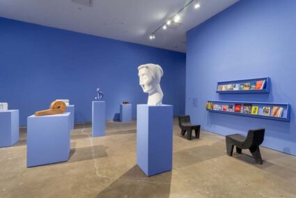 Installation view of exhibition by Pedro Reyes at SITE Santa Fe, with blue-colored walls and pedestals with various sculptures and a shelf of multi-colored books.