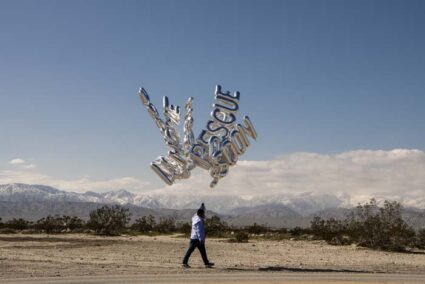 Performance by Héctor Zamora, holding silver balloons that spell out words like "Rescue", while walking around the Coachella Valley like a street vendor.
