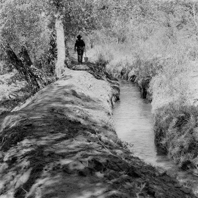Black and white photograph of an acequia with a person walking alongside it on the left side.