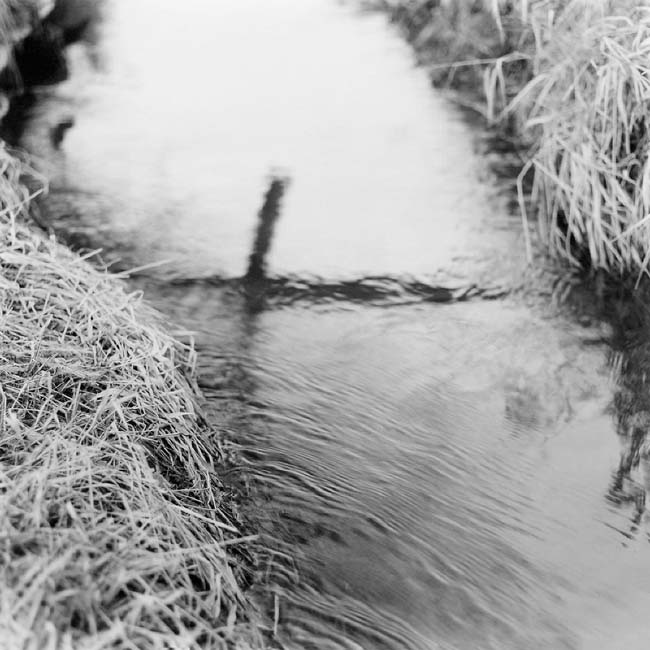 Black and white photograph of a cross reflected in the water flowing through an acequia.