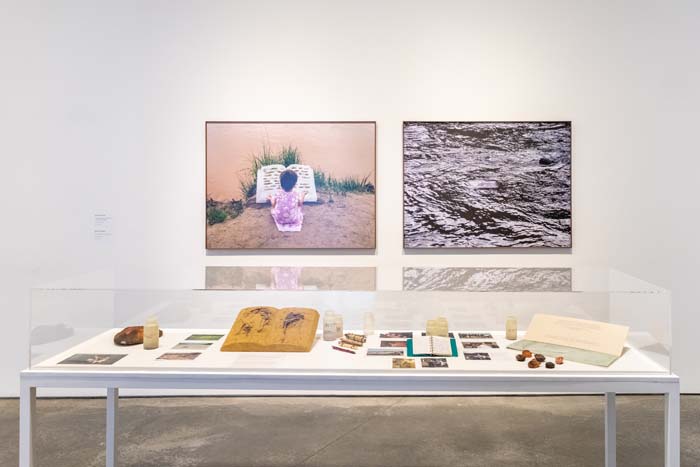 Installation view of Going With the Flow, with work by Basia Irland, with two large-scale photographs in the background, and a vitrine with objects in the foreground.
