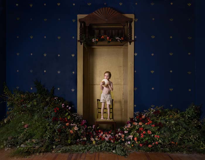 A child in a doorway in a room with blue walls and flowering bushes spread out in front of him