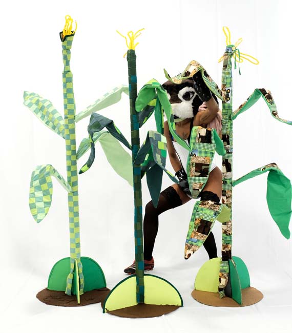 Three soft sculpture cornstalks partially obscure a figure wearing a plush cow head and lingerie