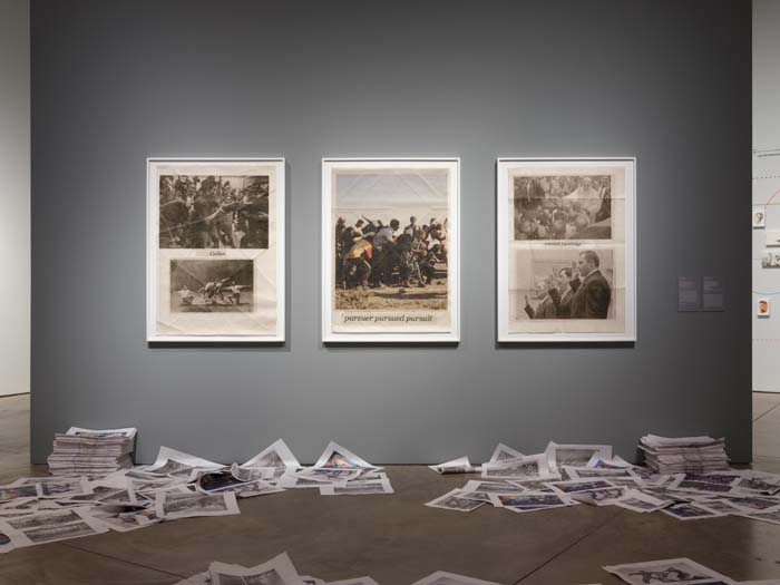 Three newspaper broadsheets with images of current events hang on the wall surrounded by stacked and scattered newspapers on the floor beneath