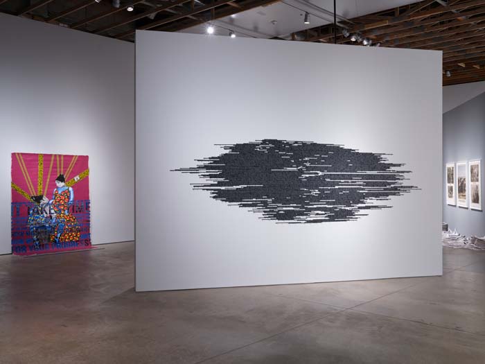 Installation view with a cloud shaped form of dense black lettering on a white wall, with a colorful painting behind it