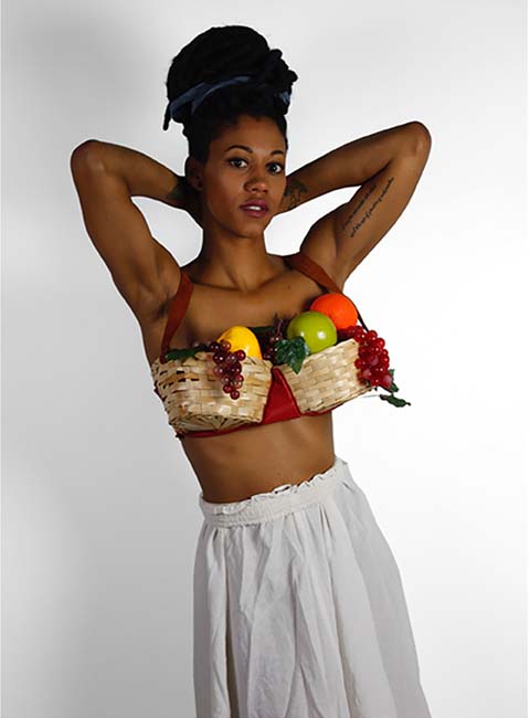 A Black woman wearing a top made of baskets overflowing with fruit, her hands tucked behind her head