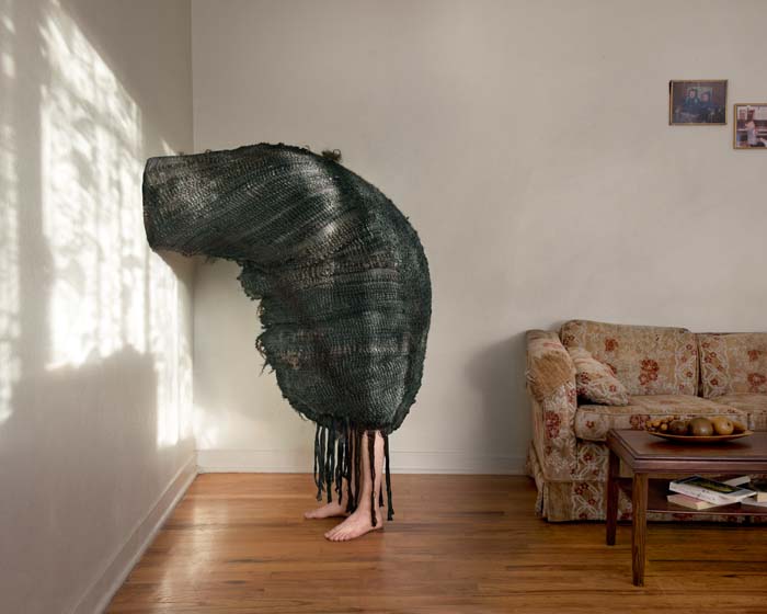 A figure standing with head resting on a wall ensconced in a shroud made of hair that obscures the body except for the feet