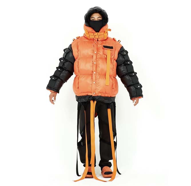 A figure wearing an oversized orange puffer jacket partially obscuring the face with straps of black and orange fabric hanging from below