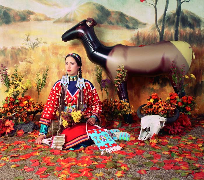Photograph of a woman in Native American dress seated in front of a painted backdrop, with an inflatable deer, a bison skull, and silk fall leaves scattered on the ground.
