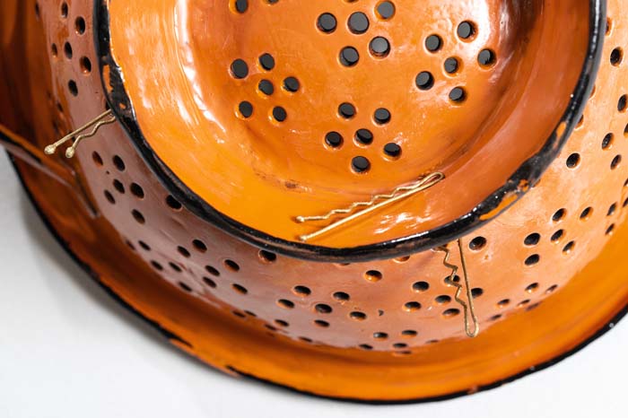Detail view of an orange painted colander with blonde bobby pins strewn about on it.