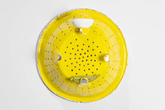 Sculpture by Tamara Johnson of a bright yellow colander hung on a wall with deviled egg and okra resting on the legs.