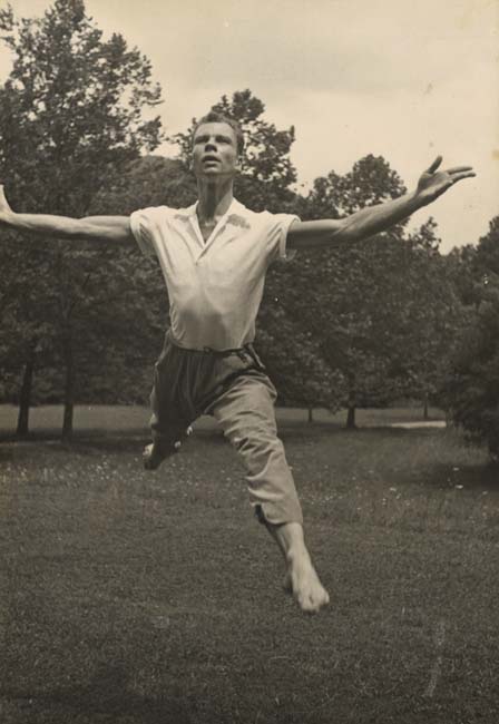 Black and white photograph of Merce Cunningham leaping with arms outstretched in a grassy field with trees in the background.