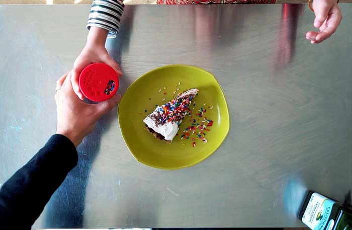 An overhead view looking down at a slice of cake with rainbow sprinkles on it and two hands reaching over a table grasping a bottle of sprinkles.