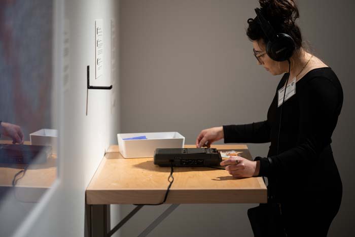 Woman wearing black headphones listening to a tape player that is on a wooden shelf.