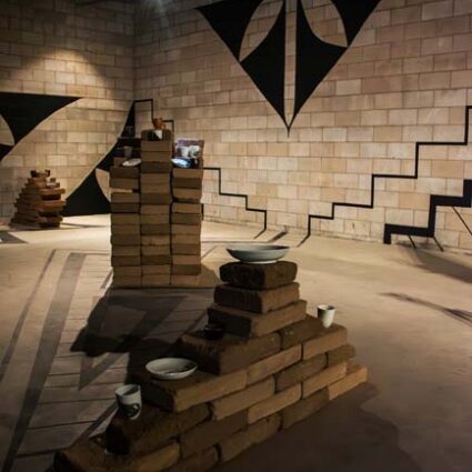 Installation view of a room lined with adobe brick and festooned with black geometric patterns, with brick pedestals holding ceramic works by artist Margarita Paz-Pedro.