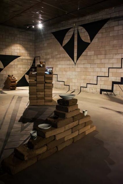 Installation view of a room lined with adobe brick and festooned with black geometric patterns, with brick pedestals holding ceramic works by artist Margarita Paz-Pedro.