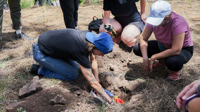 Eco art by artist Kaitlin Bryson in a blue ball cap and black t-shirt burying some multi-colored fabric in the dirt with onlookers.