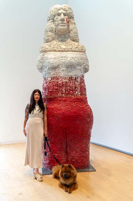 Artist Raven Halfmoon, wearing a long white dress, with her dog, standing next to a twelve-foot ceramic sculpture of a figure with a red lower half and white upper half.