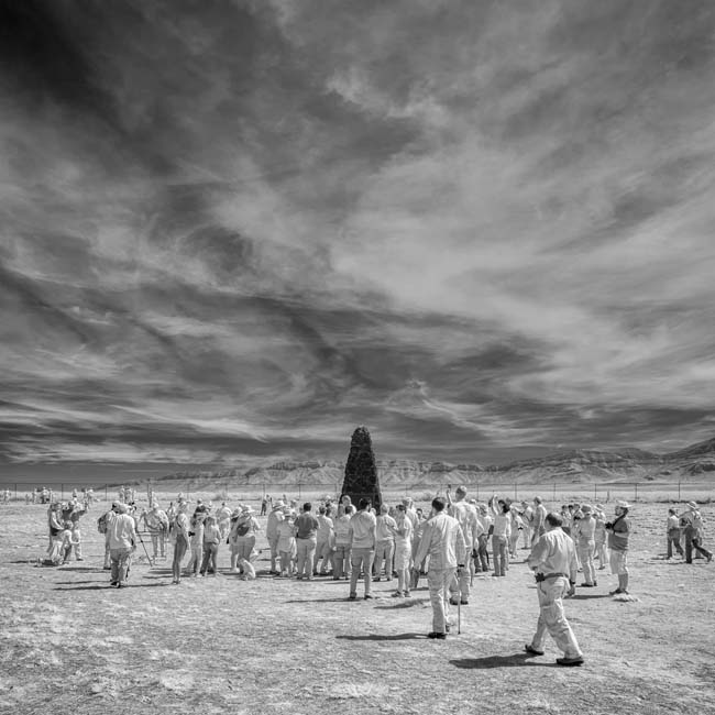 Black and white photograph showing a group of people, all seemingly dressed in white, approaching the black obelisk at the Trinity Site.