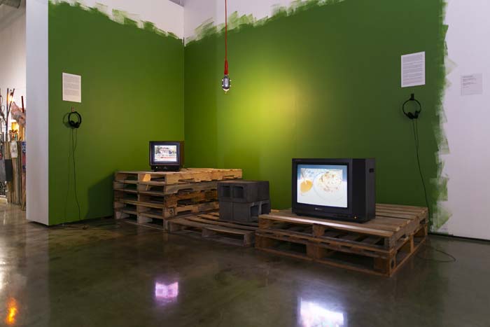 Installation view of Soy de Tejas with two vintage televisions perched on pallets, the wall behind the installation is deep green with roller marks at the edges.
