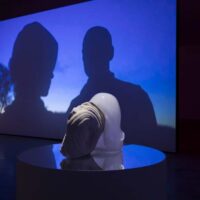 Glass sculpture by Rachel Rose set on a reflective dais in front of a screen on which two silhouetted figures appear in a twilight scene.