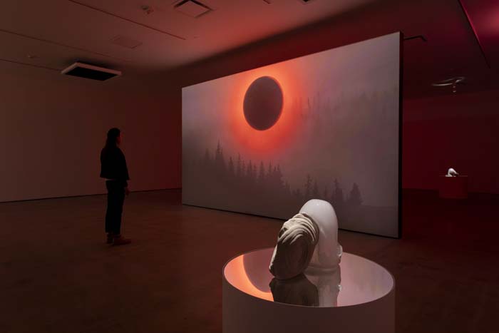 Installation view with a glass sculpture set on a reflective dais in front of a screen with a black orb radiating red rays, with a viewer standing to the left of the screen.