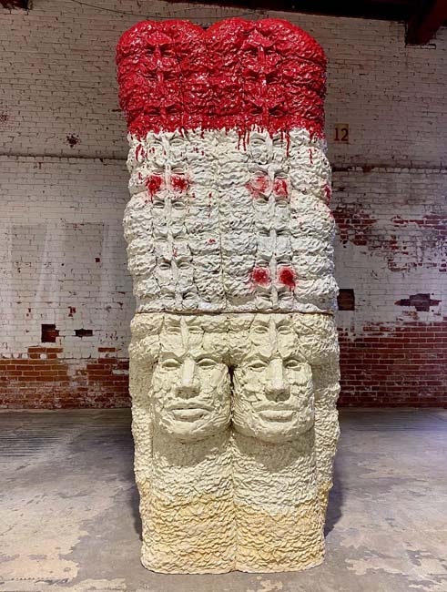 Stoneware sculpture of two heads side by side and multiple heads extending upward from them, with red paint on the top, white paint on the bottom.