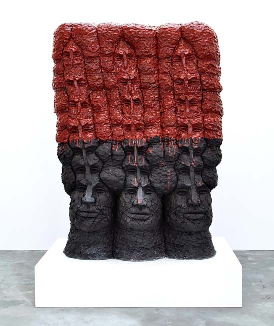 Large stoneware sculpture of three heads, with multiple heads extending above them, with the top half painted with red glaze, the bottom half in black.