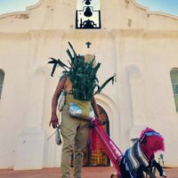 Figure in brown pants with corn stalks obscuring the face, holding a hot-pink dog-like contraption on a lead, standing in front of a mission in El Paso.