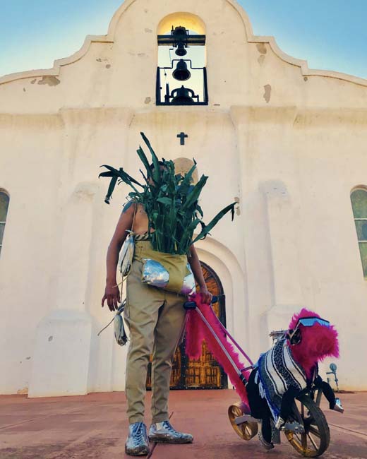 Figure in brown pants with corn stalks obscuring the face, holding a hot-pink dog-like contraption on a lead, standing in front of a mission in El Paso.