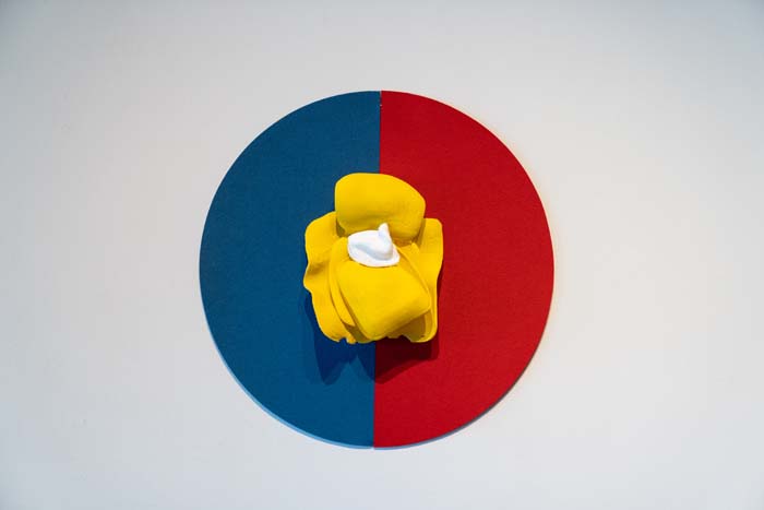Circular artwork with left half painted blue, the right half red, with a brigh yellow crumpled form with a dollop of white in the center.