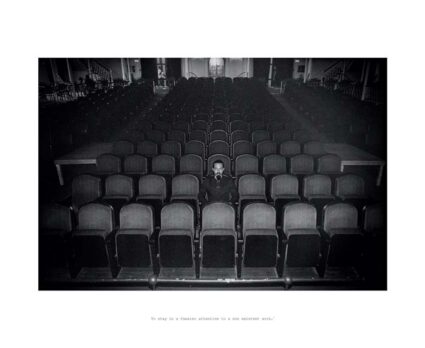 Black and white photograph of a man sitting in the middle of an otherwise empty theater with the words "To stay in a theater attentive to a non existent work" printed below.
