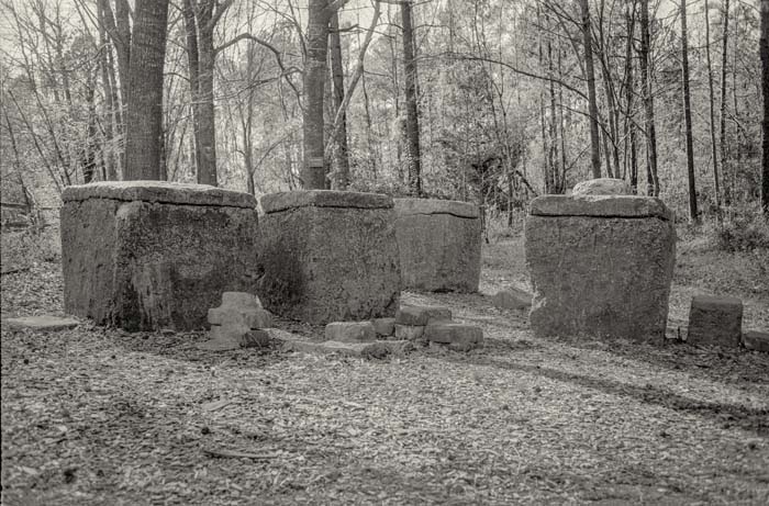Black and white photograph of a series of monumental rough hewn stones in a wooded landscape.