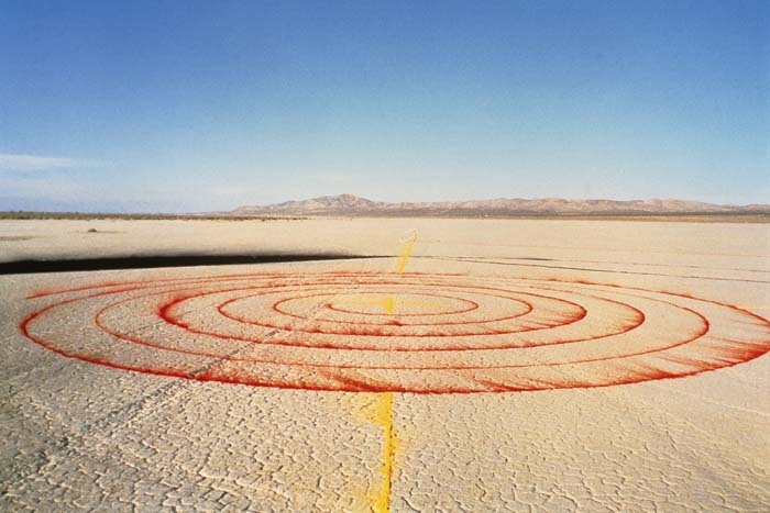 A red spiral on a dry lake bed in the desert, with a yellow line bisecting it.