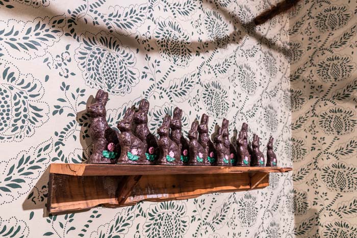 A row of chocolate bunnies on a shelf with ornate floral wallpaper behind it.