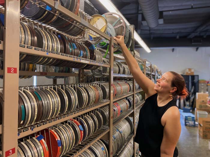 A woman reaching to the top shelf of a rack holding hundeds of film reels.