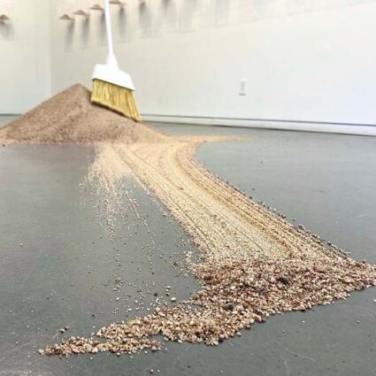 A large pile of sand on a gray floor with a broom resting on it.