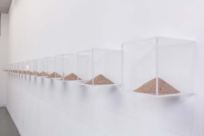 A series of Plexiglas cubes affixed to a wall, each holding a tiny pyramid-shaped pile of sand.