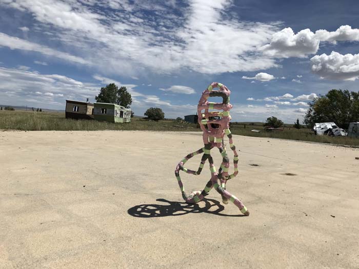 Pink and green tentacled sculpture on a concrete pad outdoors with an abandoned trailer in the background, part of the exhibition Re-Activate in the uranium mine ghost town of Shirley Basin, Wyoming.
