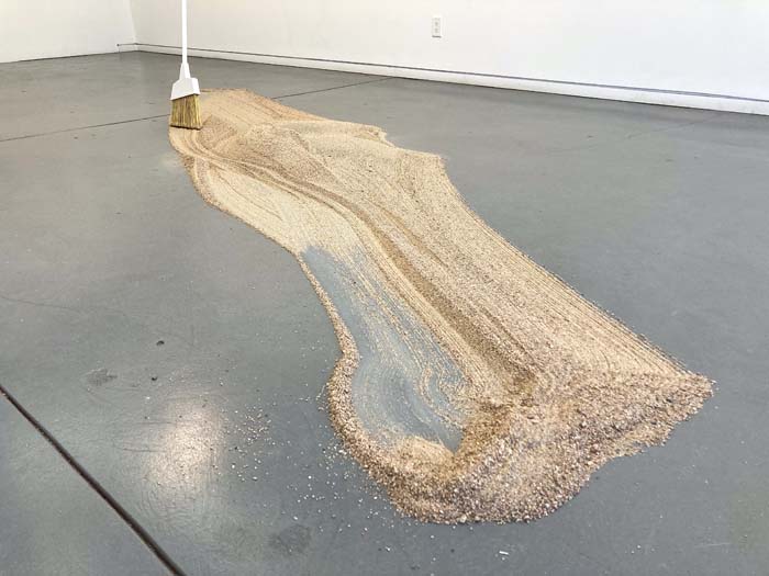 A large scattered pile of sand on a gray floor with a broom resting on it.