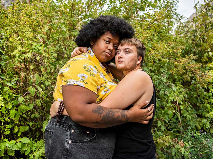 A queer couple embracing with a background of green leafy bushes.