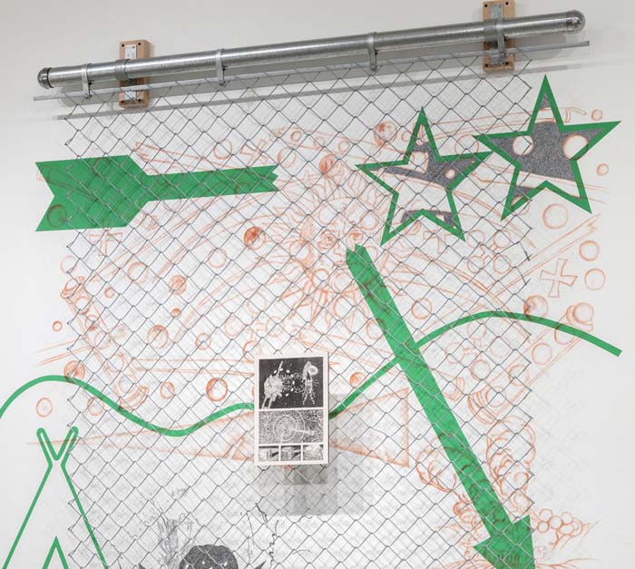 Wall piece with bright green infographic-style markings, with a broken arrow, stars, and a rudimentary landscape, with a panel of chain link suspended in front of it.