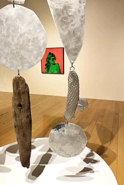 View of a hanging sculpture made of metal panels of various shapes in the foreground and a photograph with a green figure and red background in the background.