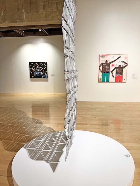 Installation view of Mythopoetica at the Palm Springs Art Museum, with a hanging sculpture comprised of metal panels casting a geometric shadow on the ground bisecting the frame.