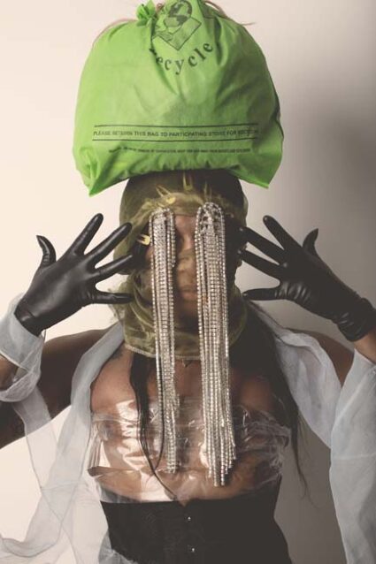 A Black femme with a green bag resting on her head and silver strands of beads covering her eyes, wearing black gloves with hands splayed near her face.