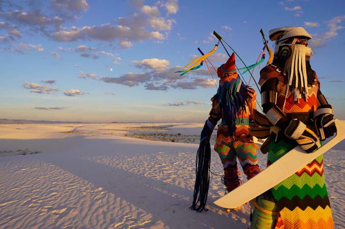 Video still of two figures in colorful crochet dress crossing a vast white desert.