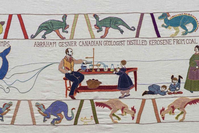 Detail of a linen tapestry with various dinosaurs depicted on the upper and lower registers, with a man and a girl in 19th century dress next to a table with test tubes on top of it, and the words "Abraham Gesner Canadian geologist distilled kerosene from coal."