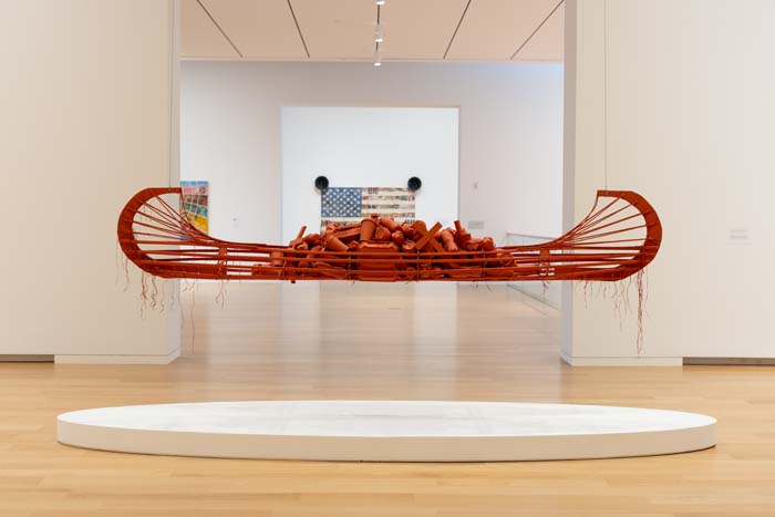 A red-colored canoe made of pinewood lath, plastic water bottles, styrofoam and paper coffee cups and take-out containers, wooden crosses, hypodermic needles, acrylic, and synthetic sinew, suspended above a dais in a museum.