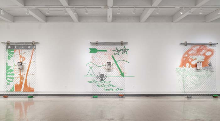 Installation view with three large-scale wall pieces comprised of chain link panels interspersed with lithograph prints and green arrow markings.